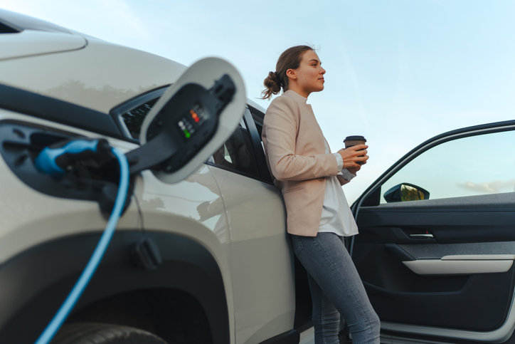 Young woman with cup of coffee waiting while her electric car charging, sustainable and economic transportation concept.